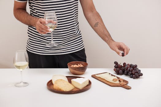 Homme en marinière tenant un verre de vin, entouré de fromages, raisins et noix sur une table blanche.