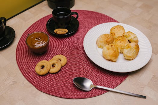 Assiette de petits pains dorés, pot de chocolat, biscuits et tasses de café sur une nappe rouge.