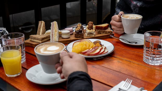 Deux personnes dégustent des pâtisseries et des boissons sur une table en bois, avec des fruits et des desserts variés.